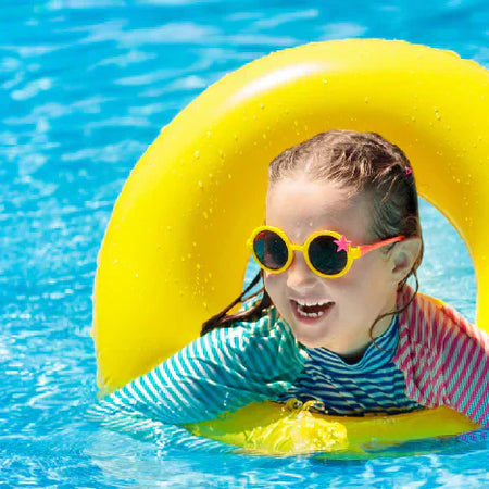 A cheerful young girl wearing yellow sunglasses and a colorful long-sleeve rash guard floating happily inside a bright yellow inner tube ring in clear, vibrant blue pool water.