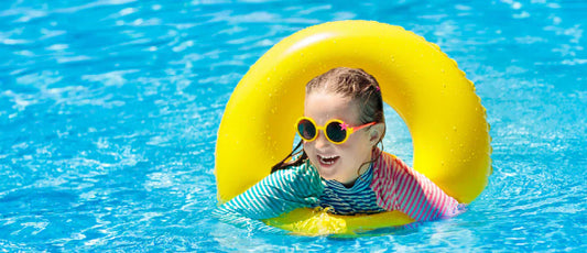 A cheerful young girl wearing yellow sunglasses and a colorful long-sleeve rash guard floating happily inside a bright yellow inner tube ring in clear, vibrant blue pool water.