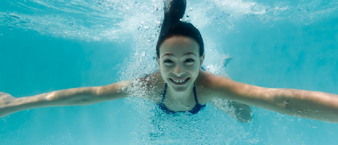 A smiling young girl in a blue swimsuit swimming smoothly underwater directly toward the camera, leaving a light trail of bubbles in crystal-clear blue pool water.