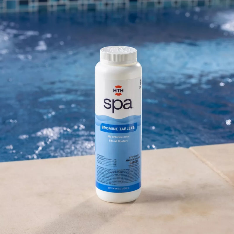 A white bottle of HTH spa Bromine Tablets sanitizer sitting on a concrete spa ledge, with turbulent blue spa water and tile background. The label displays 'BROMINE TABLETS' and details their sanitizer and no chlorine odor benefits.