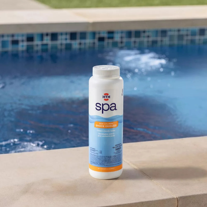 A bottle of HTH spa Non-Chlorine Shock Oxidizer sitting on a concrete spa coping stone, with turbulent blue water and mosaic tiles behind it. The orange label text features 'NON-CHLORINE SHOCK OXIDIZER' and benefits like 'Clears cloudy water'.