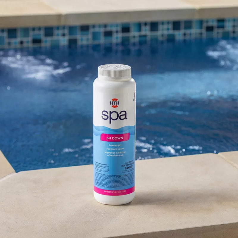 A white bottle of HTH spa pH Down liquid chemical sitting on a concrete spa ledge, with blue spa water and tile in the background. The label is clearly visible with text detailing 'pH Down', 'Lowers pH', and 'Prevents scale'.